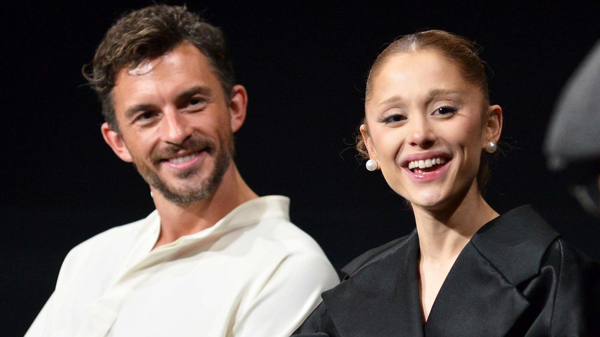 Jonathan Bailey, wearing a white open-necked shirt, and Ariana Grande in a black top and pearl stud earrings, at a Q&A on the film Wicked: For Good in Los Angeles. They are both laughing.