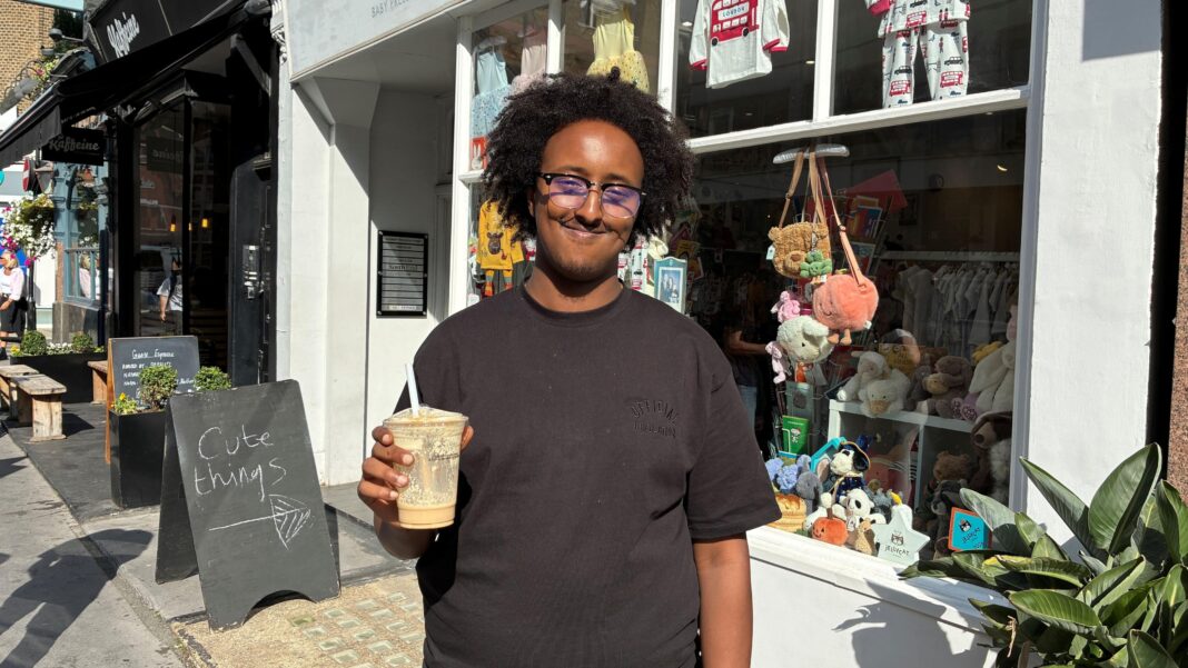 Hanad, wearing a glasses and a black T-shirt and holding an iced coffee, smiles as he stands outside a shop in London.