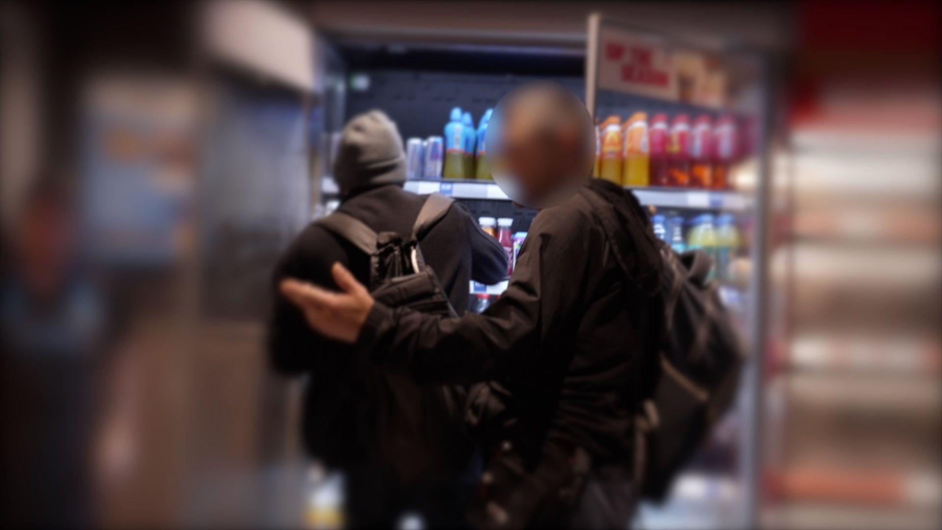 Two men wearing dark clothers and rucksacks standing in front of an open fridge in Greggs bakery. One man is jestering to a staff worker.