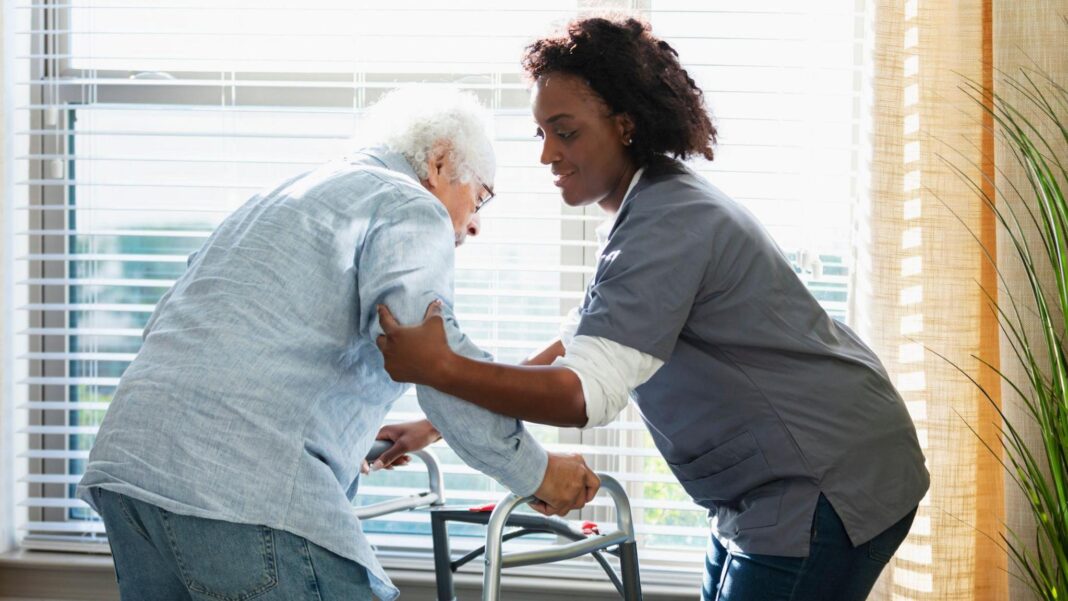 A female care home worker, wearing nursing uniform, helps support an elderly man leaning on a walking frame.