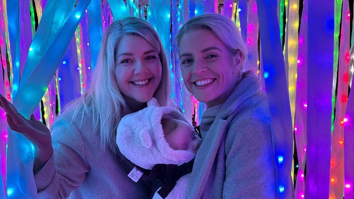 Two smiling, blonde, white women, wearing fluffy coats, standing amongst different coloured falling ribbons and fairy lights. A baby, dressed in a fluffy romper, is in a baby carried on the front of one of the women while he sleeps. There is a deep blue tinge to the photo.