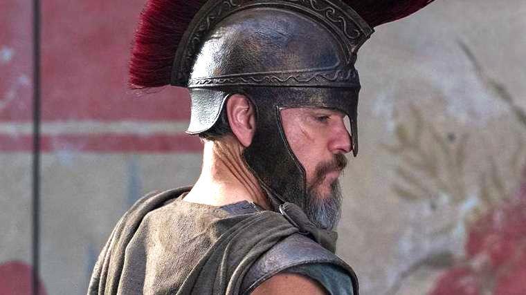 Matt Damon as Odysseus. He has a short, brown and white beard, while wearing an old-style helmet with a central plume and grey armour over his shoulders.