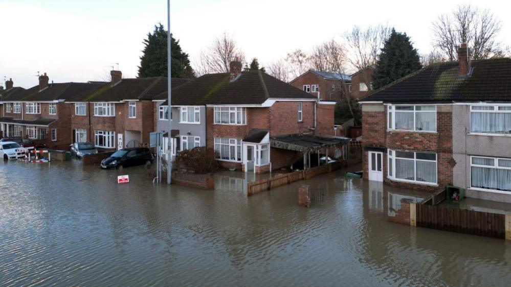 Red brick semi detached homes sit on a street that is completely underwater, with water reaching doorways but below ground floor windows.