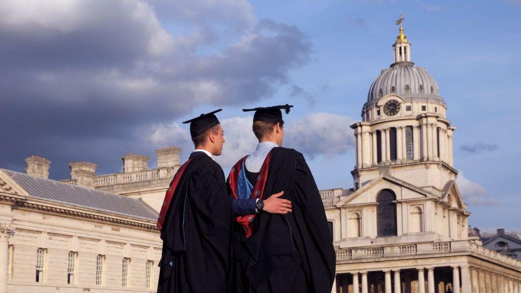 Two university graduates in black gowns and mortarboards stand with their backs to the camera, looking towards the grand, classical dome and stone buildings of the Old Royal Naval College under a cloudy sky.