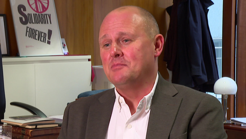 TUC general secretary Paul Nowak sitting in his office. He is pictured in a grey suit and white shirt, worn without a tie. Behind him is a desk and a poster with the phrase Solidarity Forever! on it.