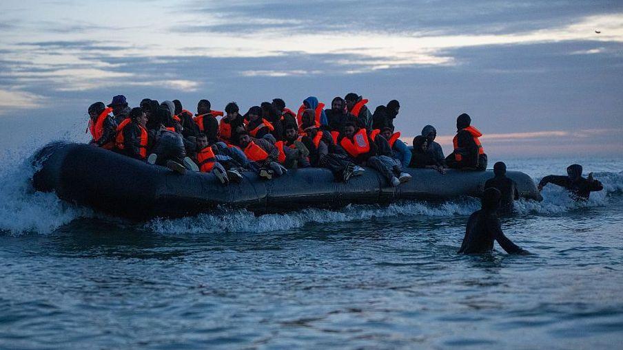 Wide shot of dozens of men crowded in orange life vests on a dinghy preparing to cross the English Channel.