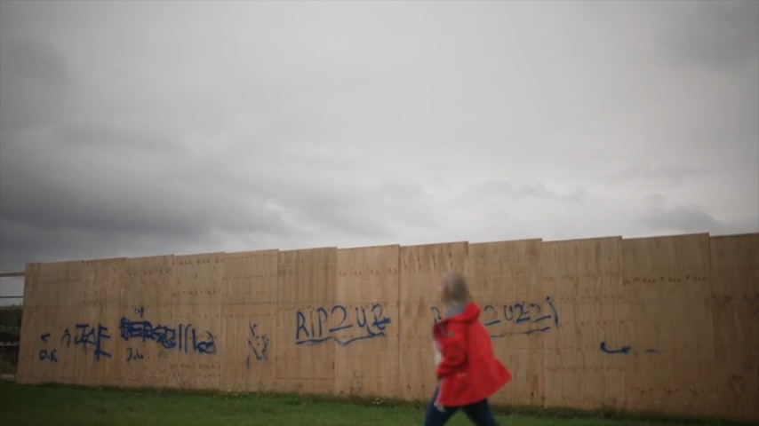 A woman in a red coat walks past some wooden board with graffiti saying RIP2UZZI