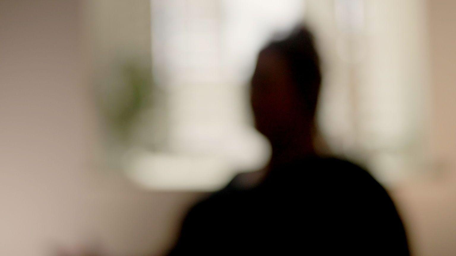 A silhouette of a woman, out of focus to further protect her identity, sitting in a white room in front of a window, with touches of green around her from what may be pot plants