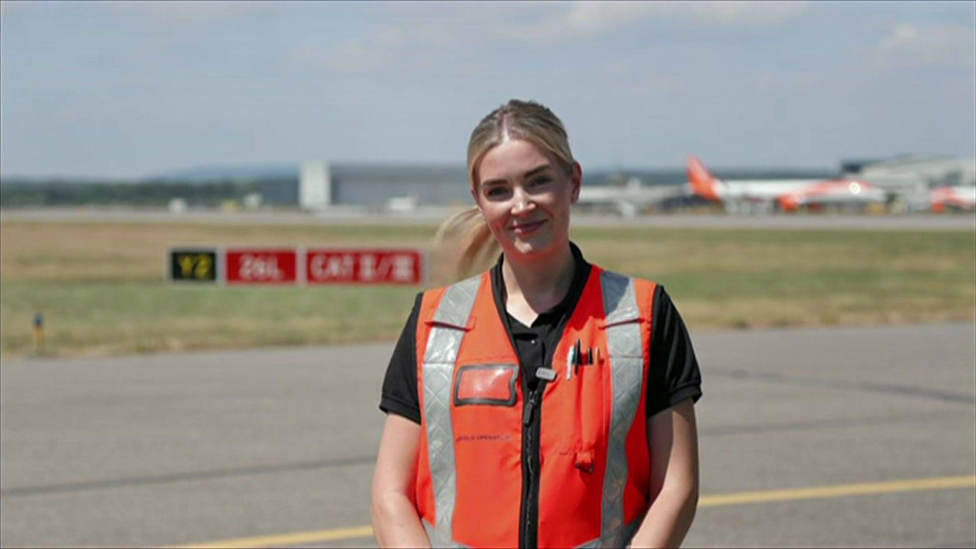Danyell stands on the side of the runway in an orange high vis jacket. Planes are in the background.
