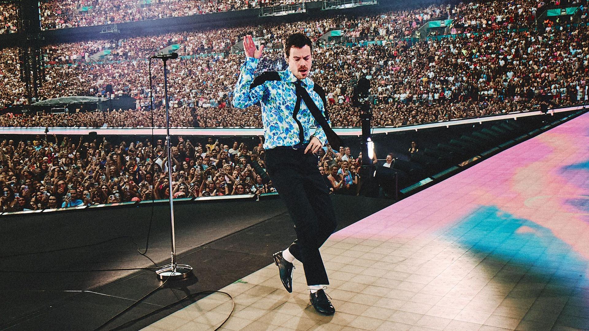 Harry Styles dancing on stage at Wembley wearing a blue floral shirt, black tie, black trousers, white socks and black patent shoes. Thousands of people can be seen standing or watching him from the stands