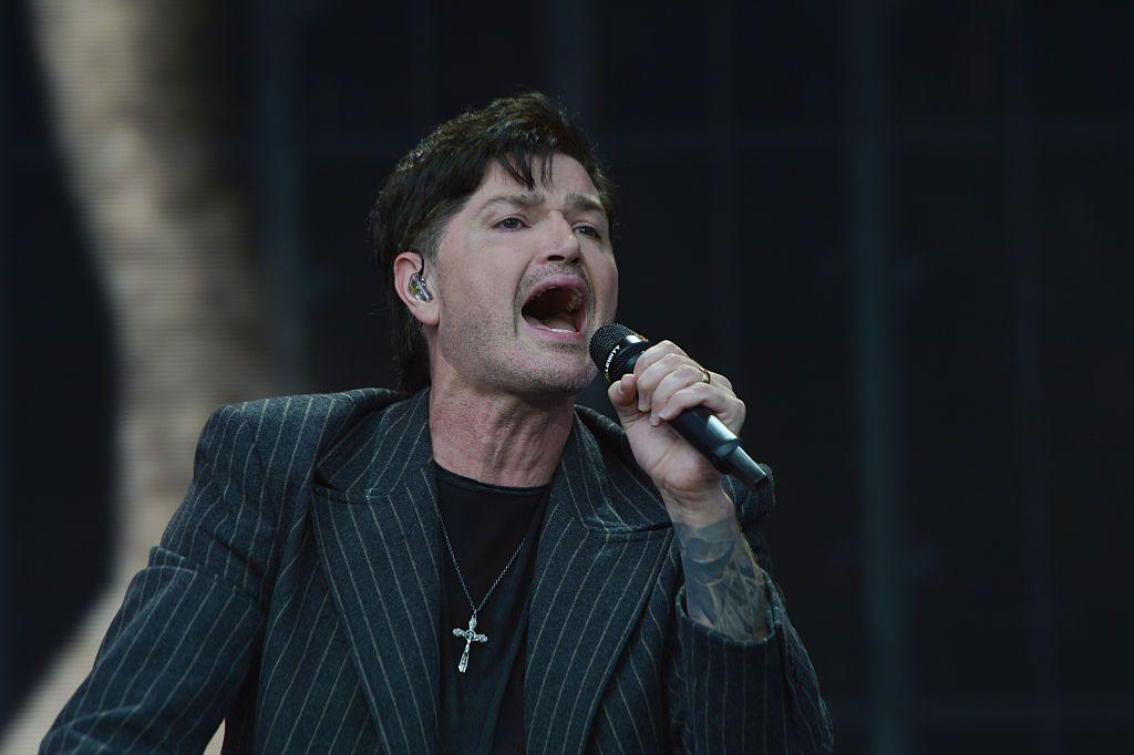 Danny O'Donoghue from The Script singing into a microphone on stage. He is wearing a black pin-striped jacket with a black top underneath. He has a large, silver cross around his neck.