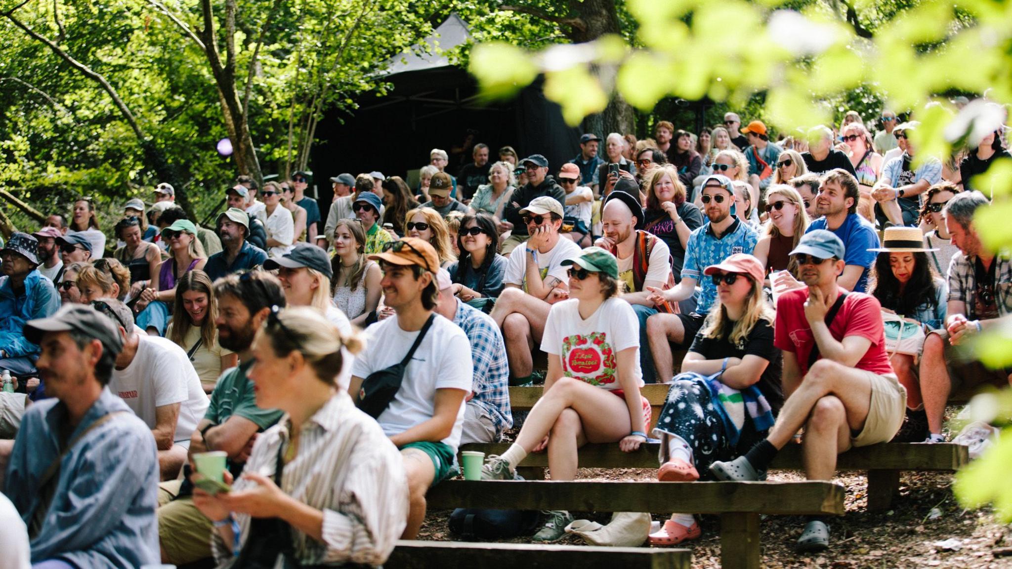People sat on benches in crowd in a forest area.