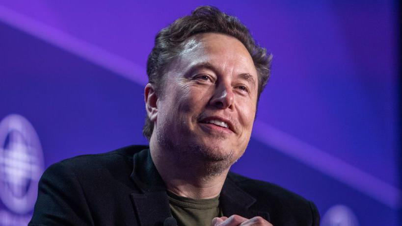 Elon Musk stares towards the camera with a smile while sitting against a purple background on a stage