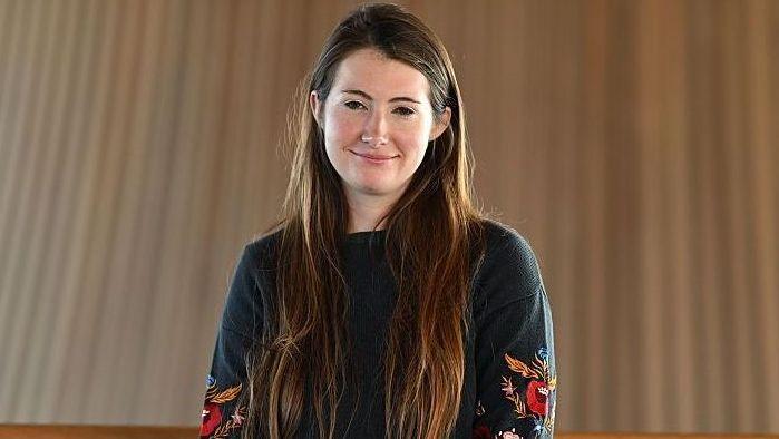 A head and shoulders shot of Sarah Cooper-Lesadd looking at the camera. She has long, straight brown hair extending below the shoulders. She is wearing a dark, long-sleeved top or sweater. Both sleeves feature colourful embroidered floral designs, including red, orange, blue, and white elements.