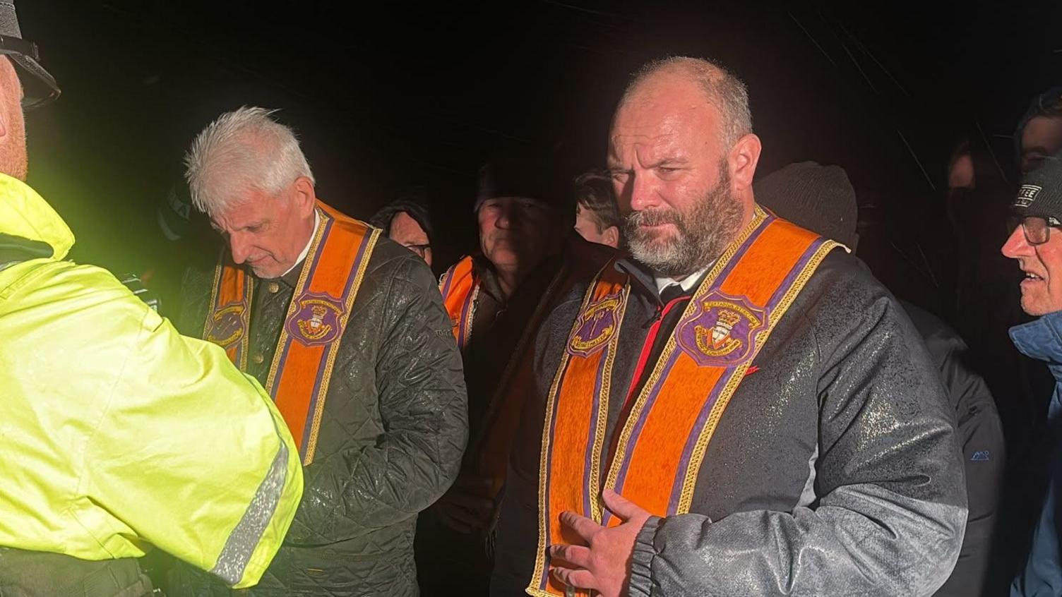 Two members of the Orange Order's Portadown District Lodge stand in front of a police officer, whose back is to the camera. It is dark outside.