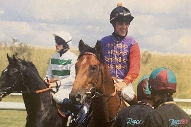 JJ Hamblett is riding a brown horse and is wearing purple and red jockey colours and a black helmet. He is talking to two people stood on his left.