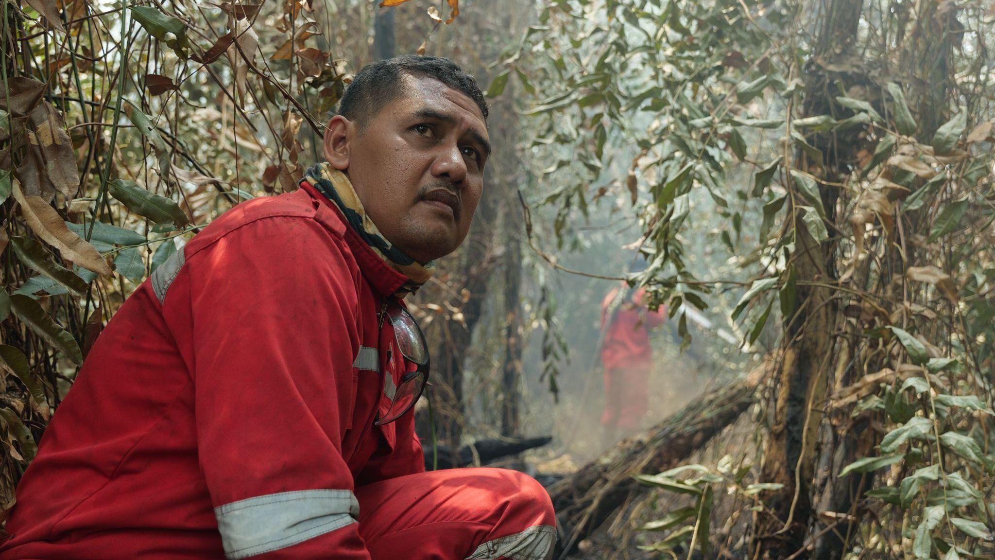 A man in red firefighting uniform crouches in bushland