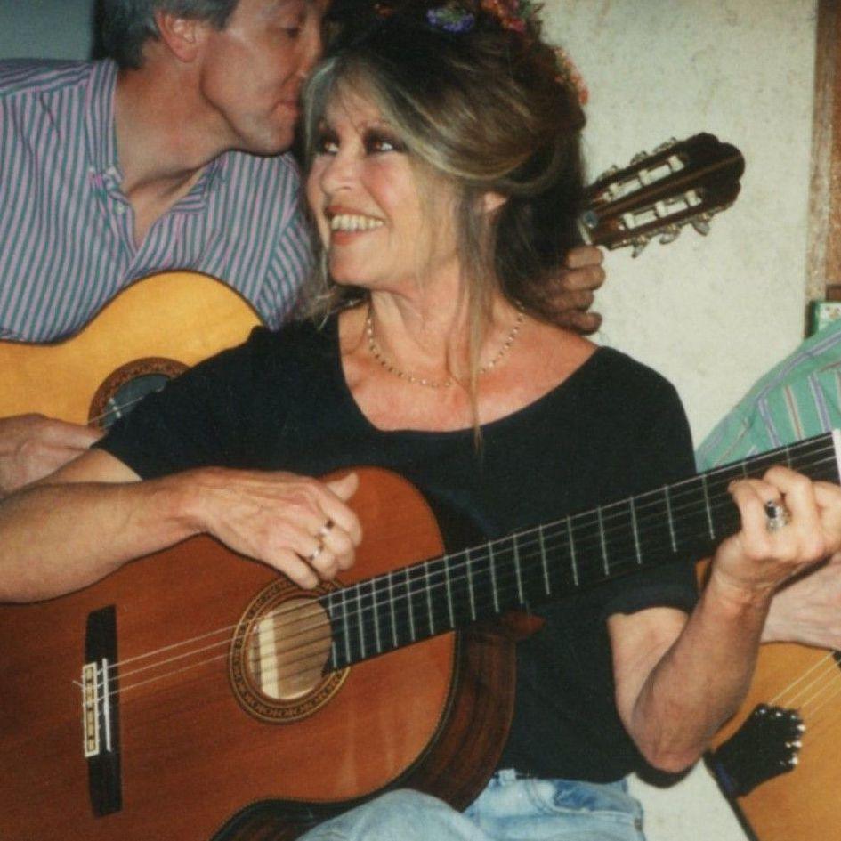Brigitte Bardot pictured playing an acoustic guitar in her later years. She has her greying hair in an up do decorated with flowers. She wears a black t-shirt and pale blue jeans.