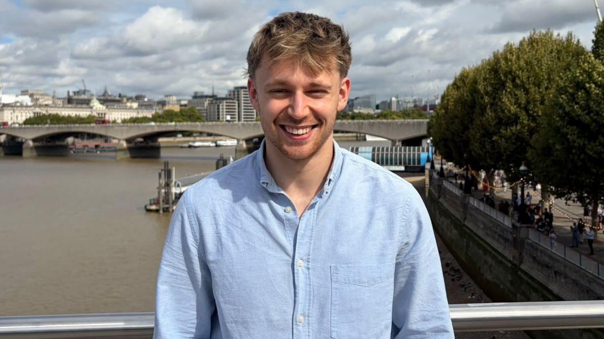 Rob is smiling at the camera. He is wearing a blue shirt. He is standing on a bridge over the river