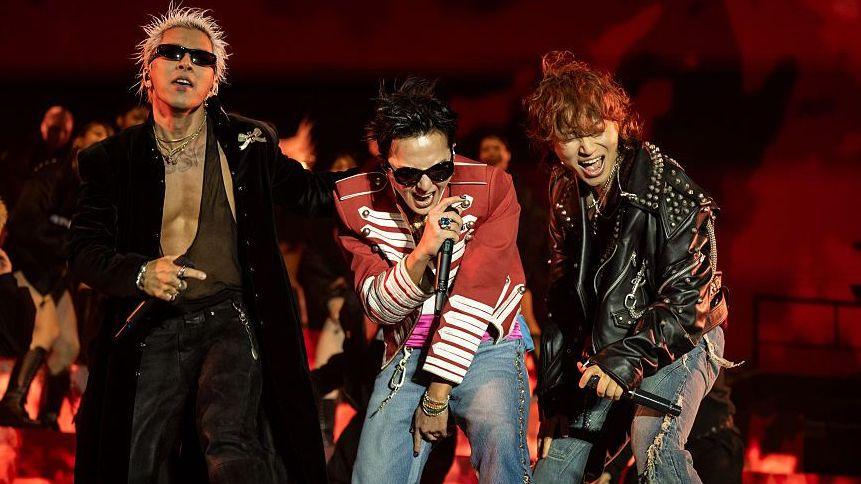 Three performers stand at the front of a stage holding handheld microphones during a live performance. One performer wears a long black coat and sunglasses, another wears a red military-style jacket with white trim and jeans, and a third wears a black studded leather jacket and ripped jeans. The stage is lit in red, with flames or flame-effect visuals displayed behind the performers.