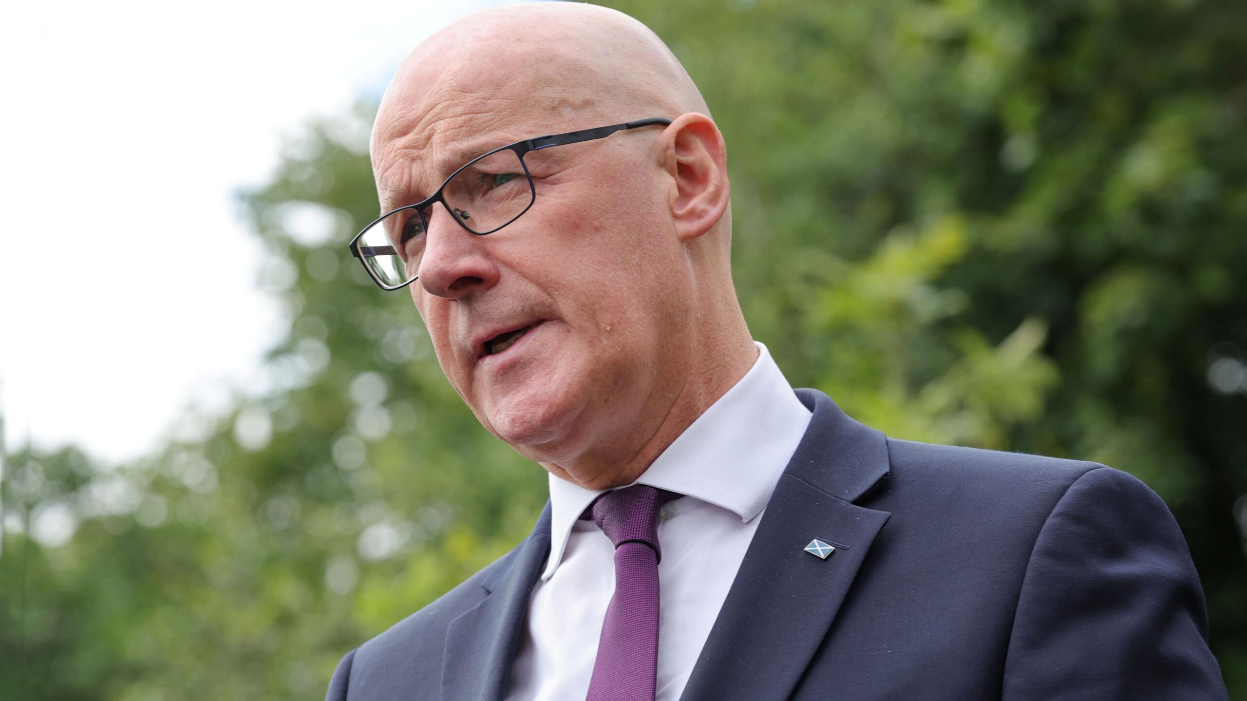John Swinney, who is bald and wearing glasses, photographed outdoors with trees in the background