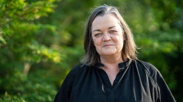A women in her 50s dressed in a black smock style shirt smiles into the camera. She is stood outdoors in front of thick green trees. She has black shoulder length hair.