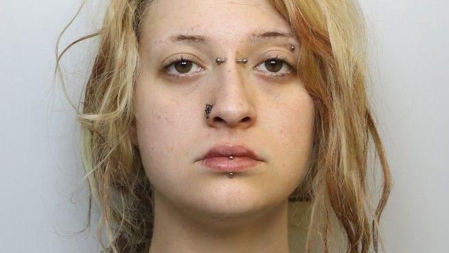 A mugshot of a young woman with blonde hair. It tied back in a messy ponytail. She has her lip, eyebrow, nose and the bridge of her nose pierced with metal bars and has a neutral expression.