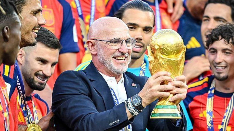 Luis de la Fuente, wearing a blue blazer, holds the gold World Cup trophy in two hands, surrounded by Spain players