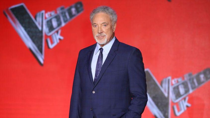 Sir Tom Jones in a blue suit stood in front of a red backdrop with the words The Voice UK on it.