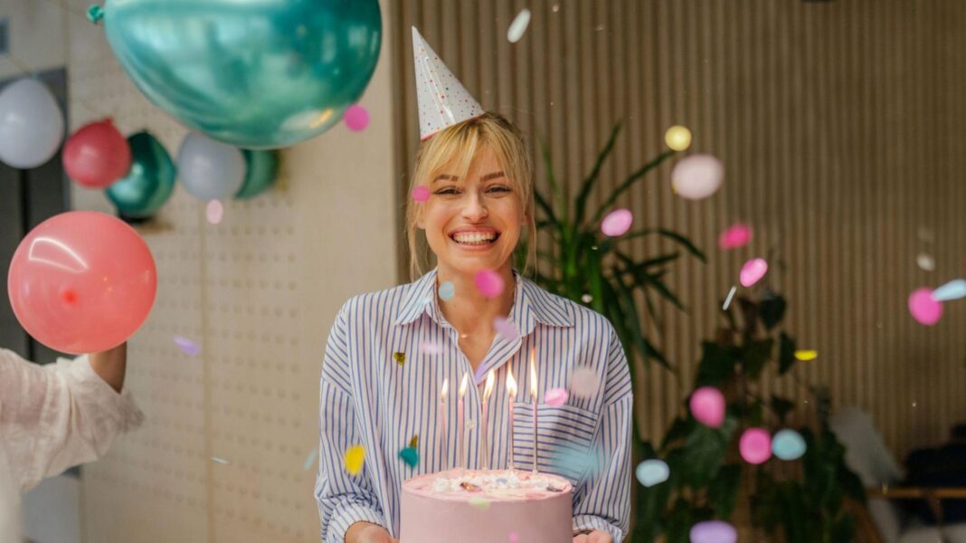 From cake to pizza: How to get the best birthday freebies (and why there’s a catch) A young woman holding a pink birthday cake with five candles on, wearing a party hat and striped shirt, celebrating her birthday, with balloons seen hanging behind her