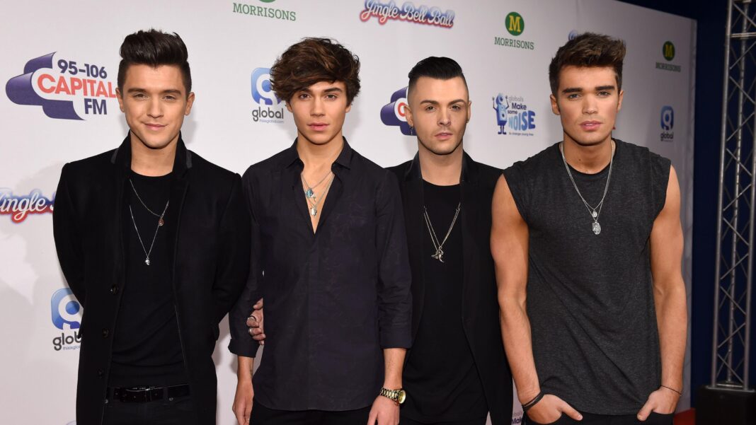 Four members of a boy band are standing side by side. They are all wearing black clothes and long necklaces. They are standing in front of a white sign.