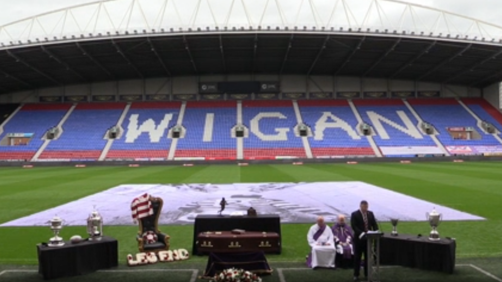 the inside of Wigan Warriors' stadium. At the bottom is the funeral. There is a table with trophies. In the middile is a coffin. On the right are two clergy sat on charis. A Man is giving a speach.