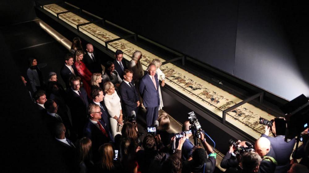 An aerial view of a group of people, with King Charles, Queen Camilla and the Macrons in the centre, standing before the Bayeux Tapestry.