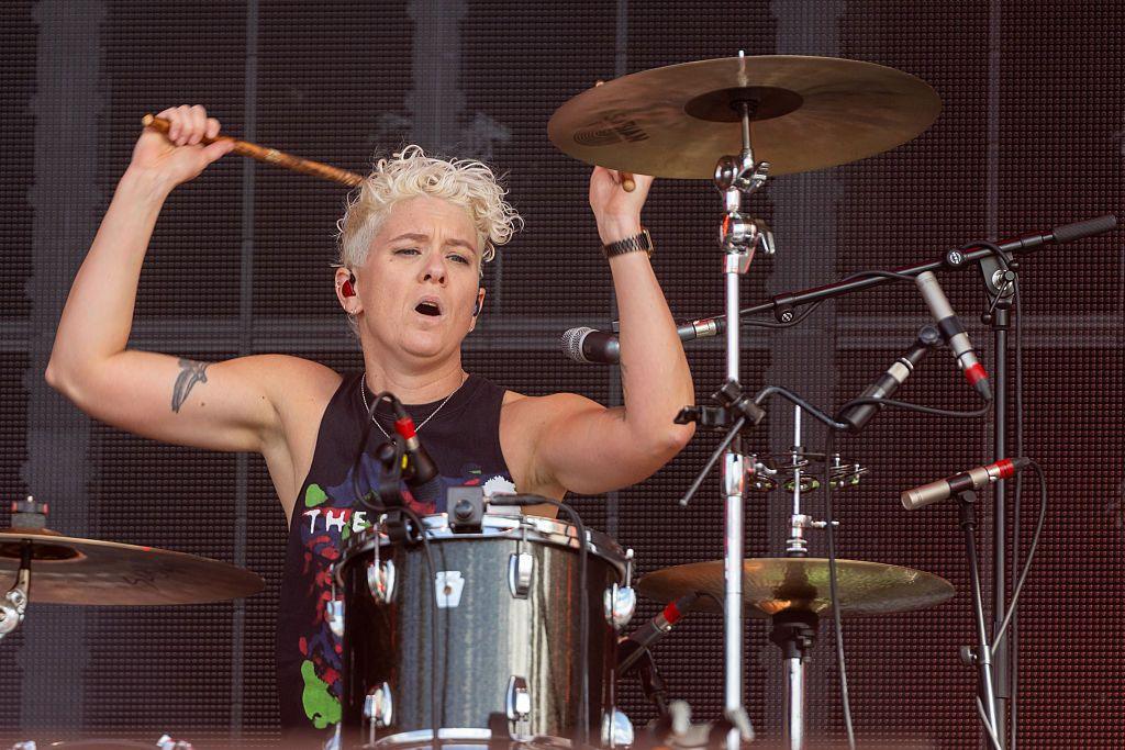 Texas drummer Cat Myers of playing on stage. She has short, curly, blonde hair and is wearing a black vest. Her arms are raised with drumsticks in her hands about to strike the drums