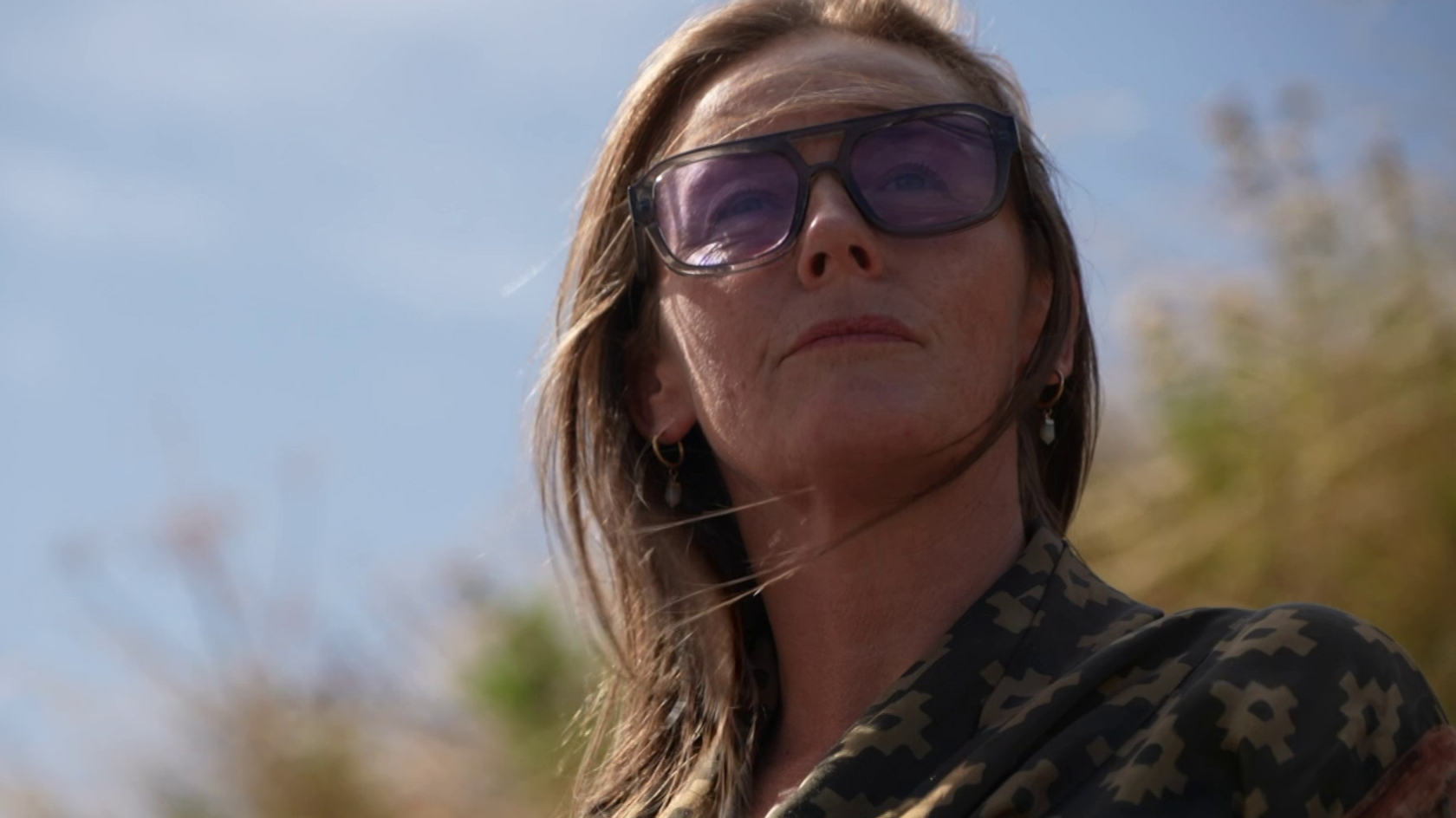 Claire Torpey standing outside looking into the distance wearing purple tinted sunglasses and her hair ius blowing slightly in the wind. She is wearing a brown scarf over her shoulders.