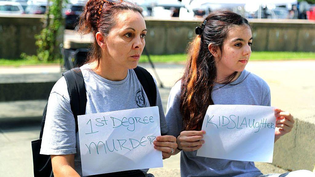 Two people with signs are seen outside the courthouse. The signs read "1st degree murderer" and "kidslaughter".