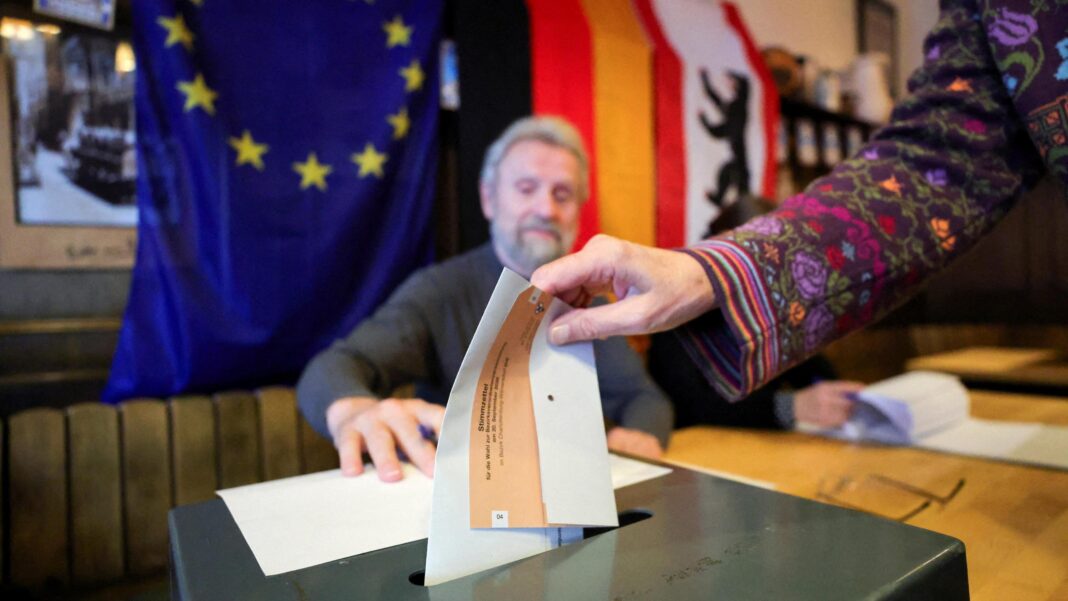 A close-up shot of a woman placing her paper vote into a black ballot box. Only her hand is visible, and the sleeve of her jumper which is purple with multicoloured flowers. A man with white hair and a beard is sat behind the desk on which the ballot box sits, and watches her deposit the vote. Behind him are the EU, German and Berlin flags.