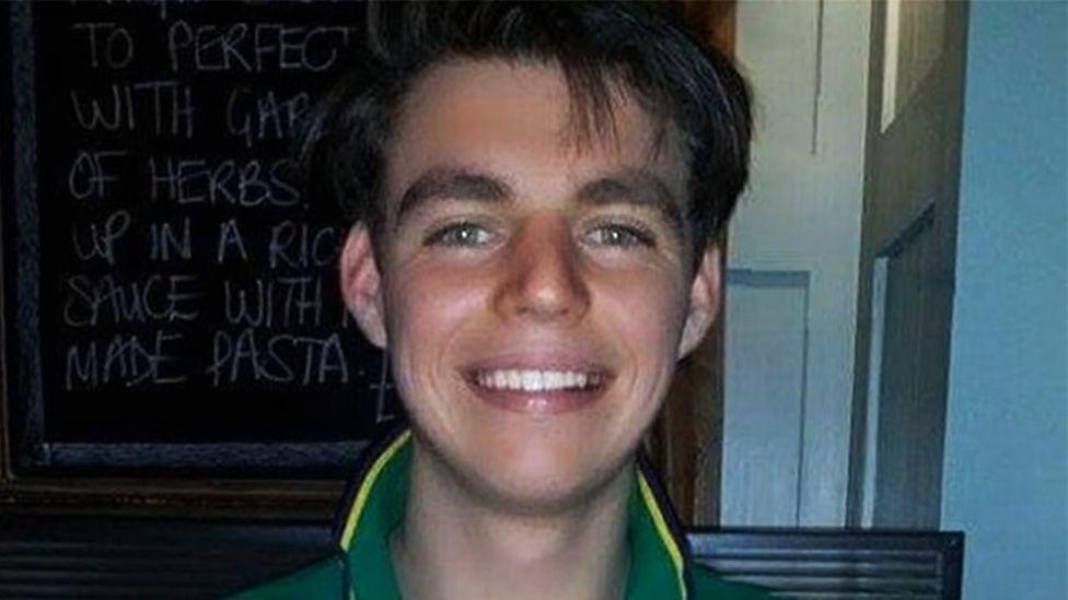 A portrait photo of Oliver McGowan who is stood in front of a blackboard in the kitchen with writing on it. He is wearing a green t-shirt and smiling with a glint in his eyes and floppy brown hair.