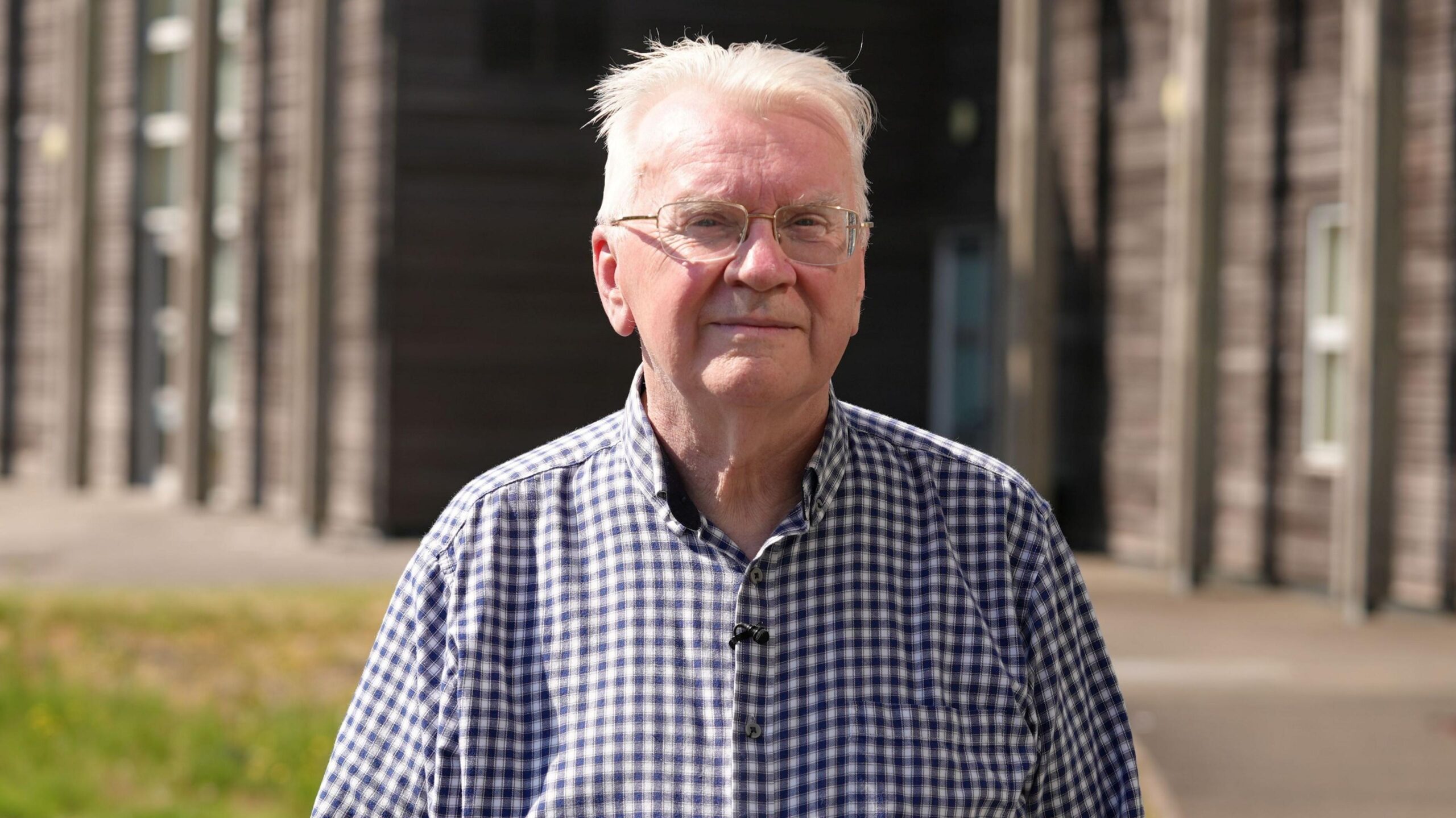 A man with white hair and a blue checked shirt looks at the camera on a sunny day