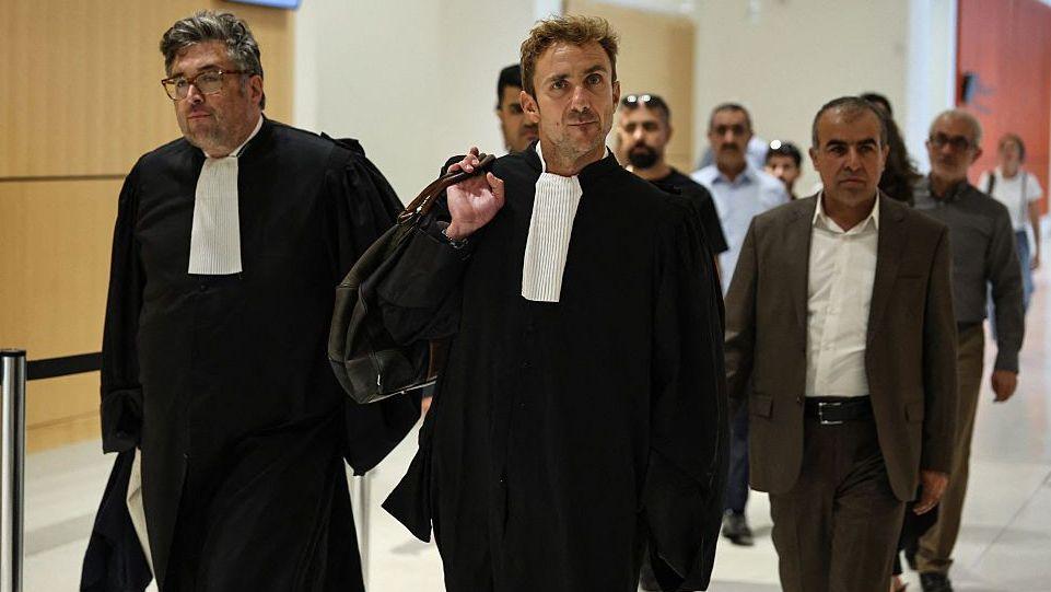 A lawyer wearing black with a white cravate strolls through a hall with other men either side