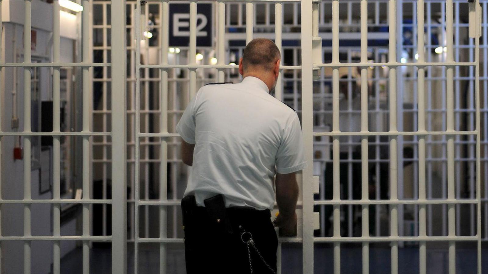 An image of a series of security gates in a prison - with floor-to-ceiling white metal bars. Above the first set it says "E/F Wing" and a prison officer - a bald man in a white shirt and black trousers - is opening the gate. We see him from behind.