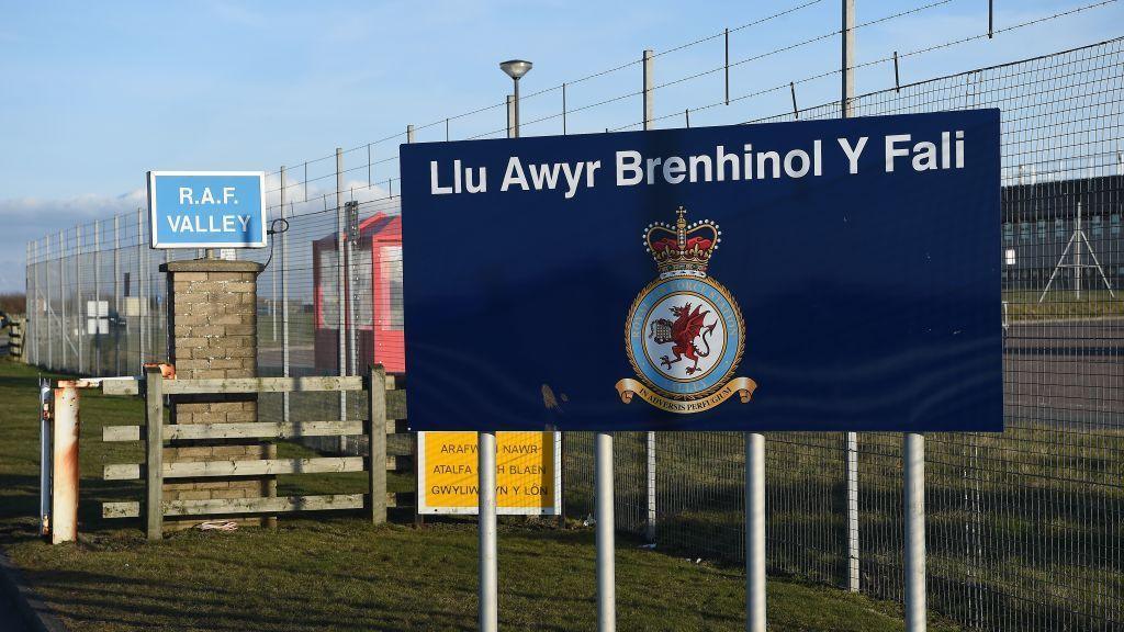 RAF Valley sign next to barbed wire fencing. A blue sign with the words Llu Awyr Brenhinol Y Fali is in the foreground.