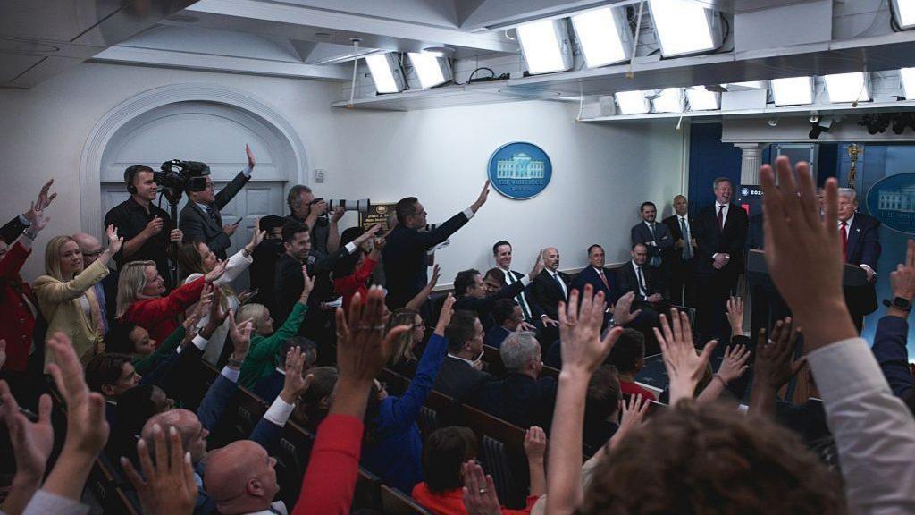 The White House press room full of reporters with their hands raised