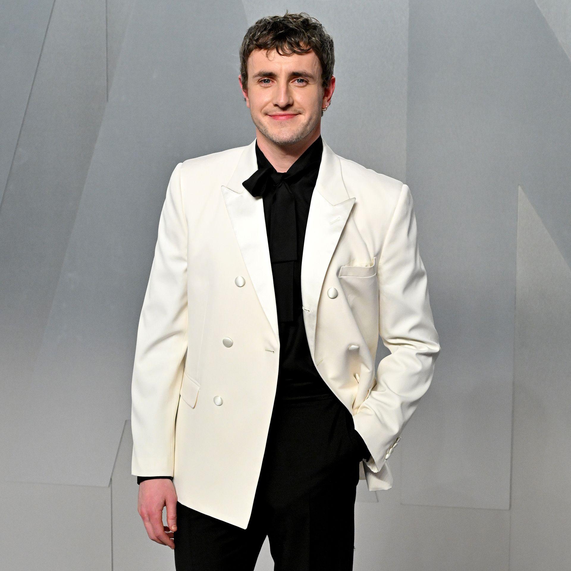 Paul Mescal smiles while posing for a photo wearing a white tuxedo with black shirt and trousers against a grey background, in LA in March.