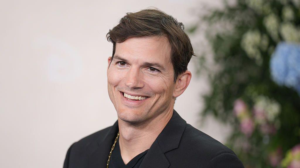 A head and shoulders shot of Ashton Kutcher in which he looks at the camera and smiles. he has on a dark jacket and a gold chain around his neck.