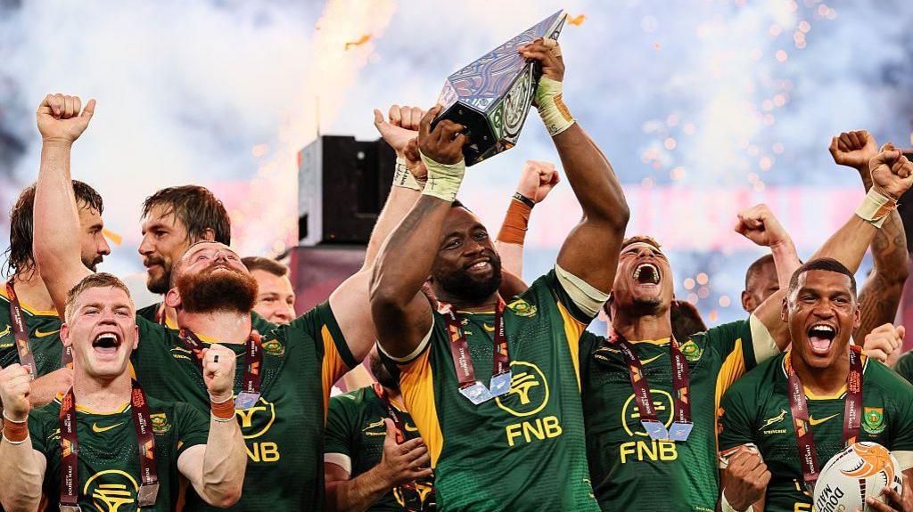 Siya Kolisi lifts the Rugby's Greatest Rivalry trophy, surrounded by happy team-mates in South Africa's largely green kit, as pyrotechnics light up the background