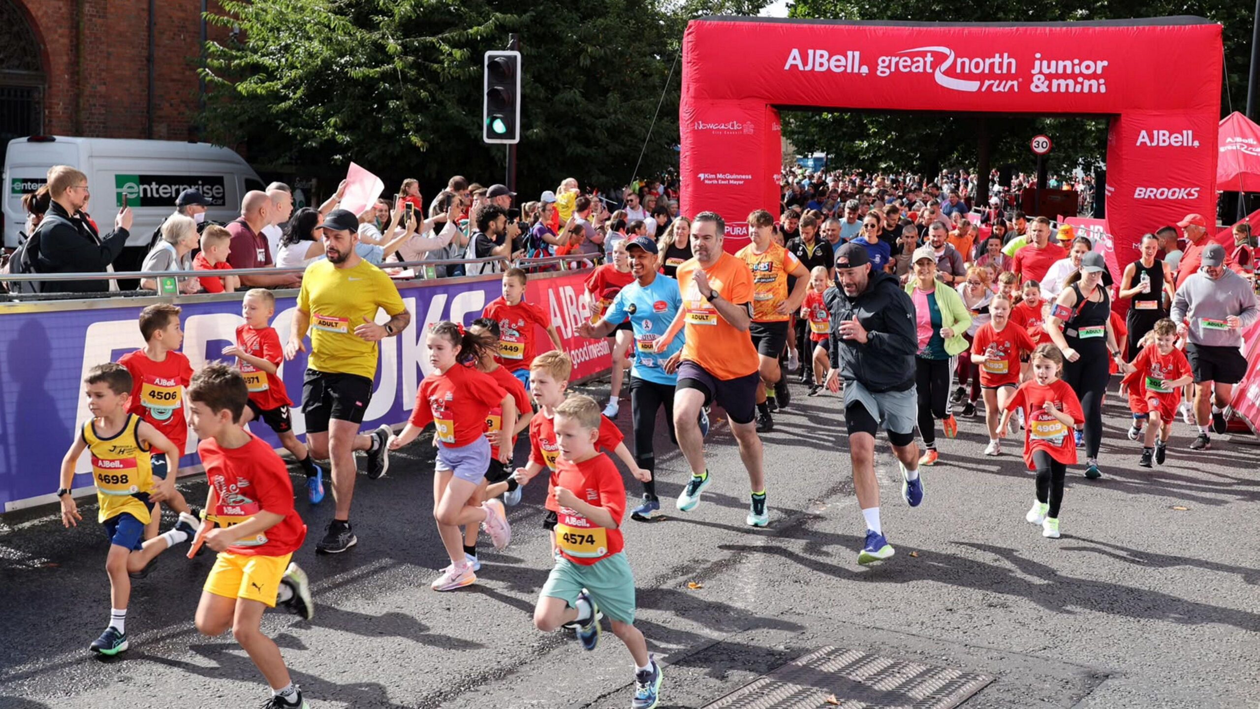 Hundreds of children are running along the cordoned off road, with adults dotted amongst them. There is an inflatable red branded archway, which reads: "AJ Bell Great North Run junior & mini". Crowds of onlookers are holding up their phones.