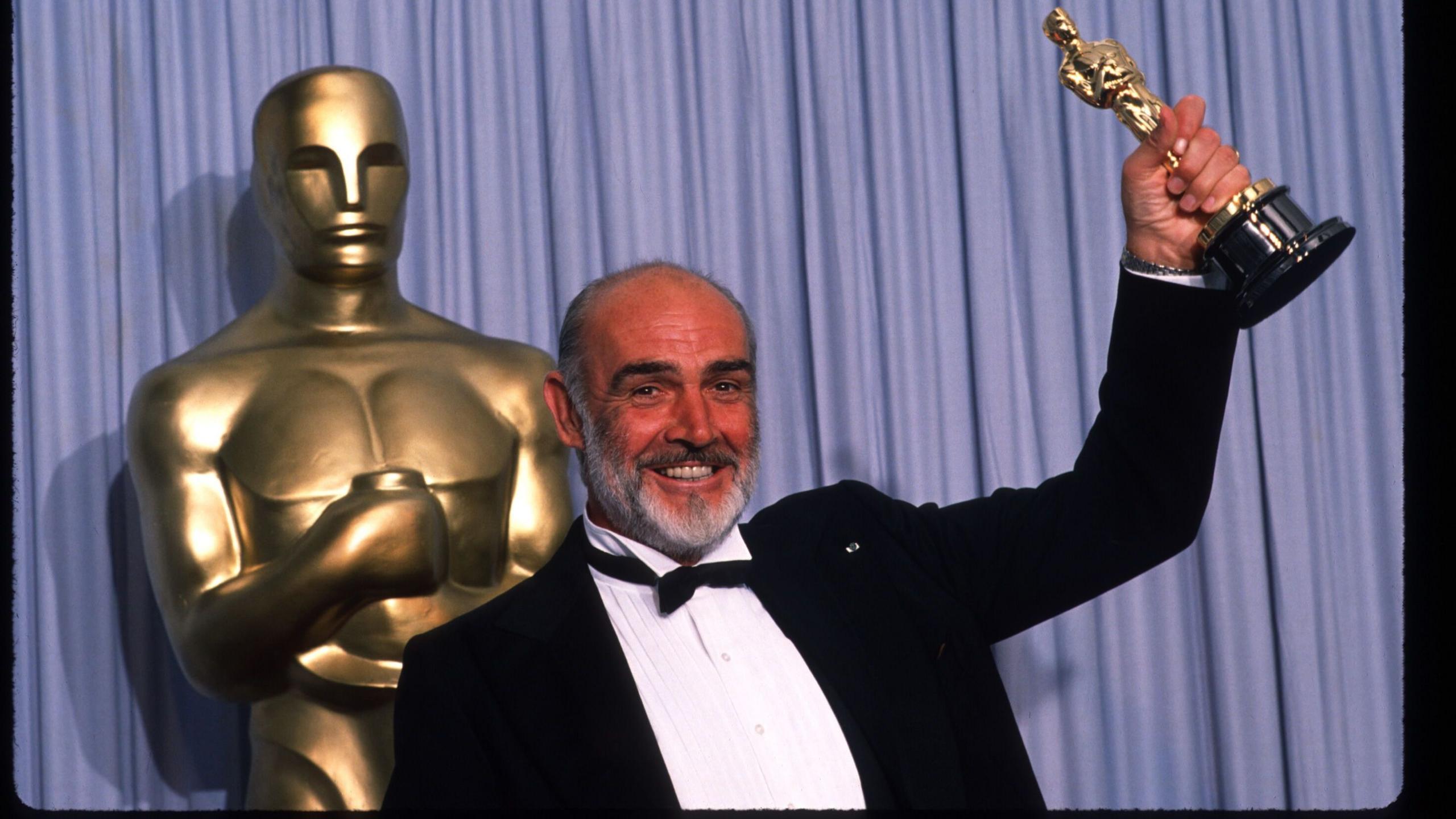 A man is in a tuxedo lift an Oscar trophy to the sky and smiling. He has grey hair and a grey beard and is stood in front of a blue curtain and oversized gold Oscar trophy.