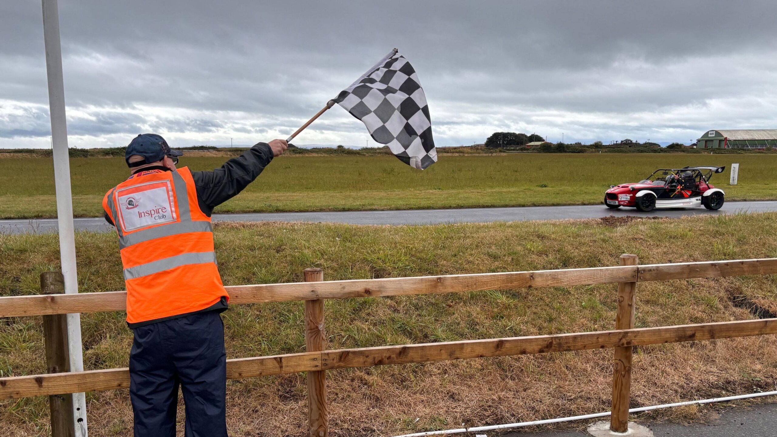 A man in a hi vis vest is holding a black and white checkered flag and waving through a red car which is on the track