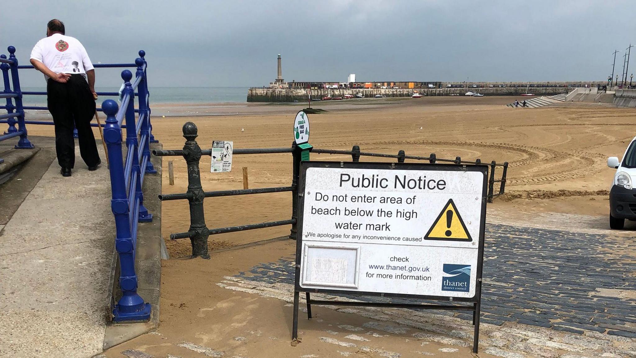 A man standing with his back to the camera at a beach. There is a warning sign urging people not to enter the water.